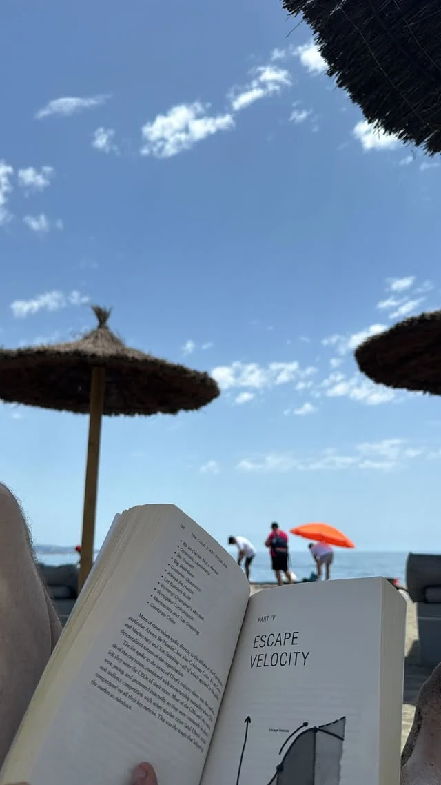 An open book beneath straw beach umbrellas, with blue sky and the sea behind it.