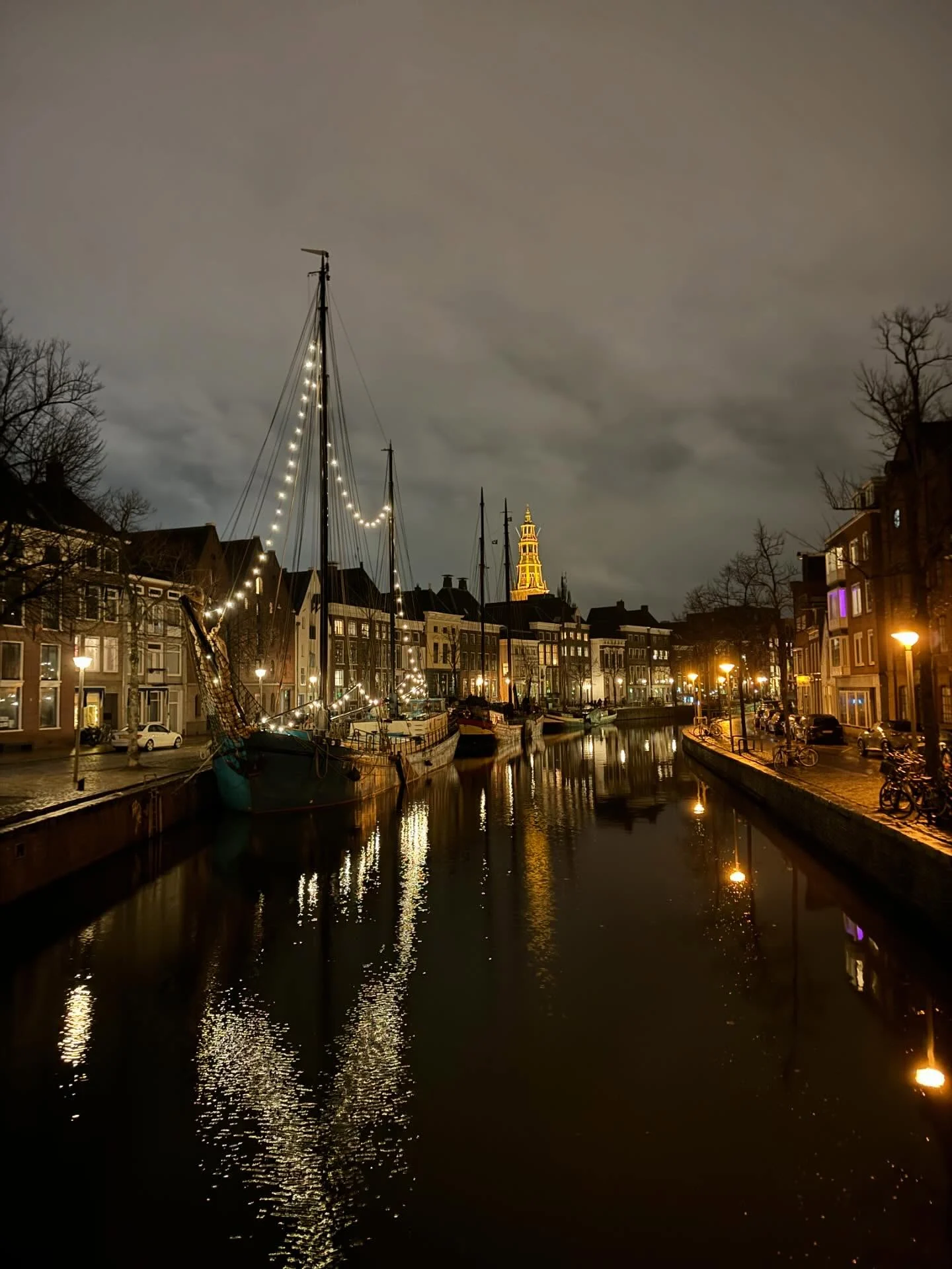 A sailboat hung with warm string lights on a quiet Dutch canal at night.