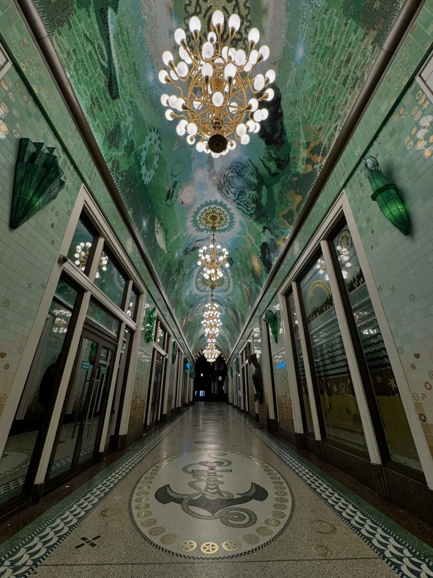 An ornate green passage with tiled walls, chandeliers and a patterned floor.