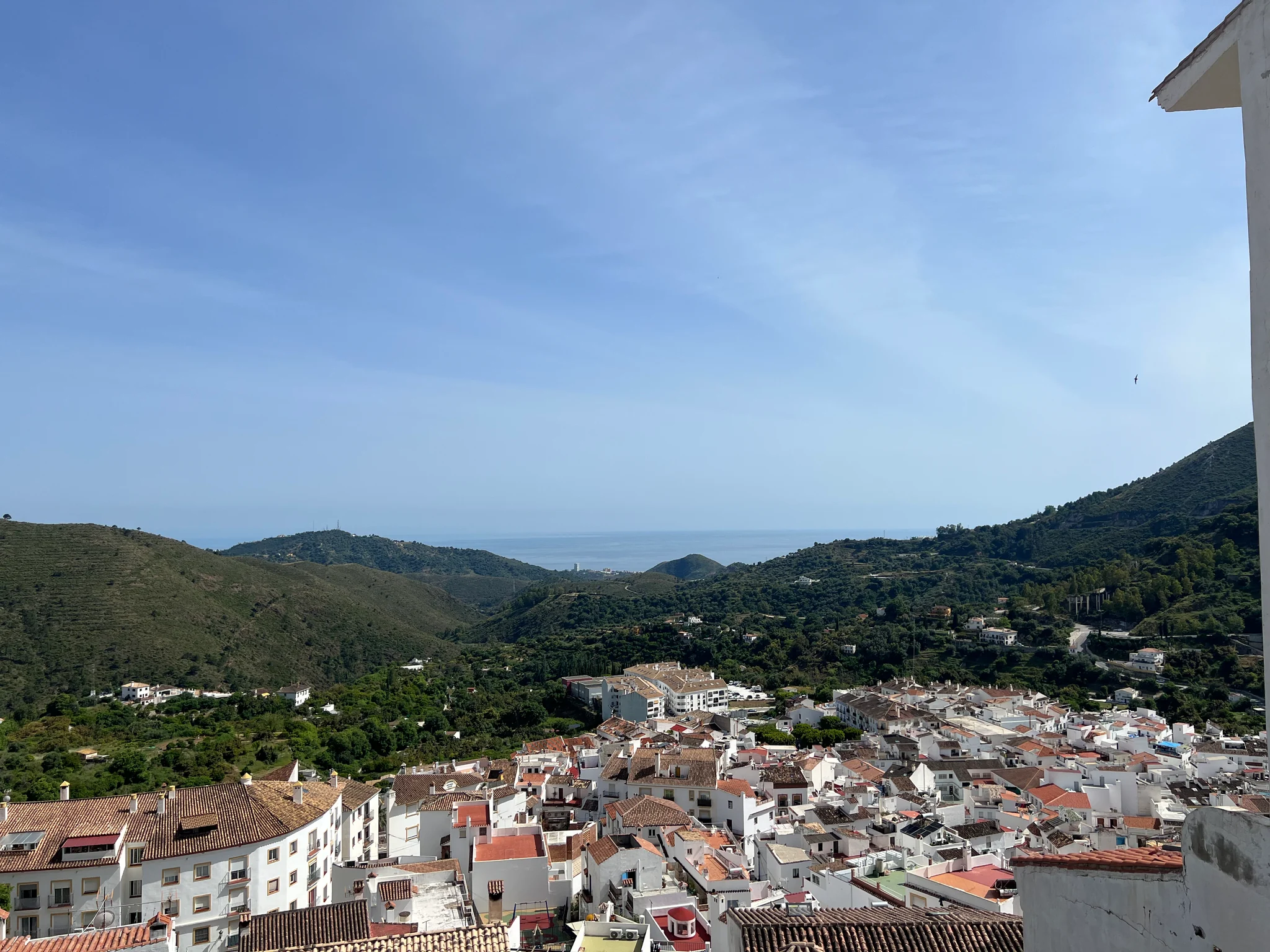 White buildings and terracotta rooftops among green hills in Málaga, beneath a pale blue sky.