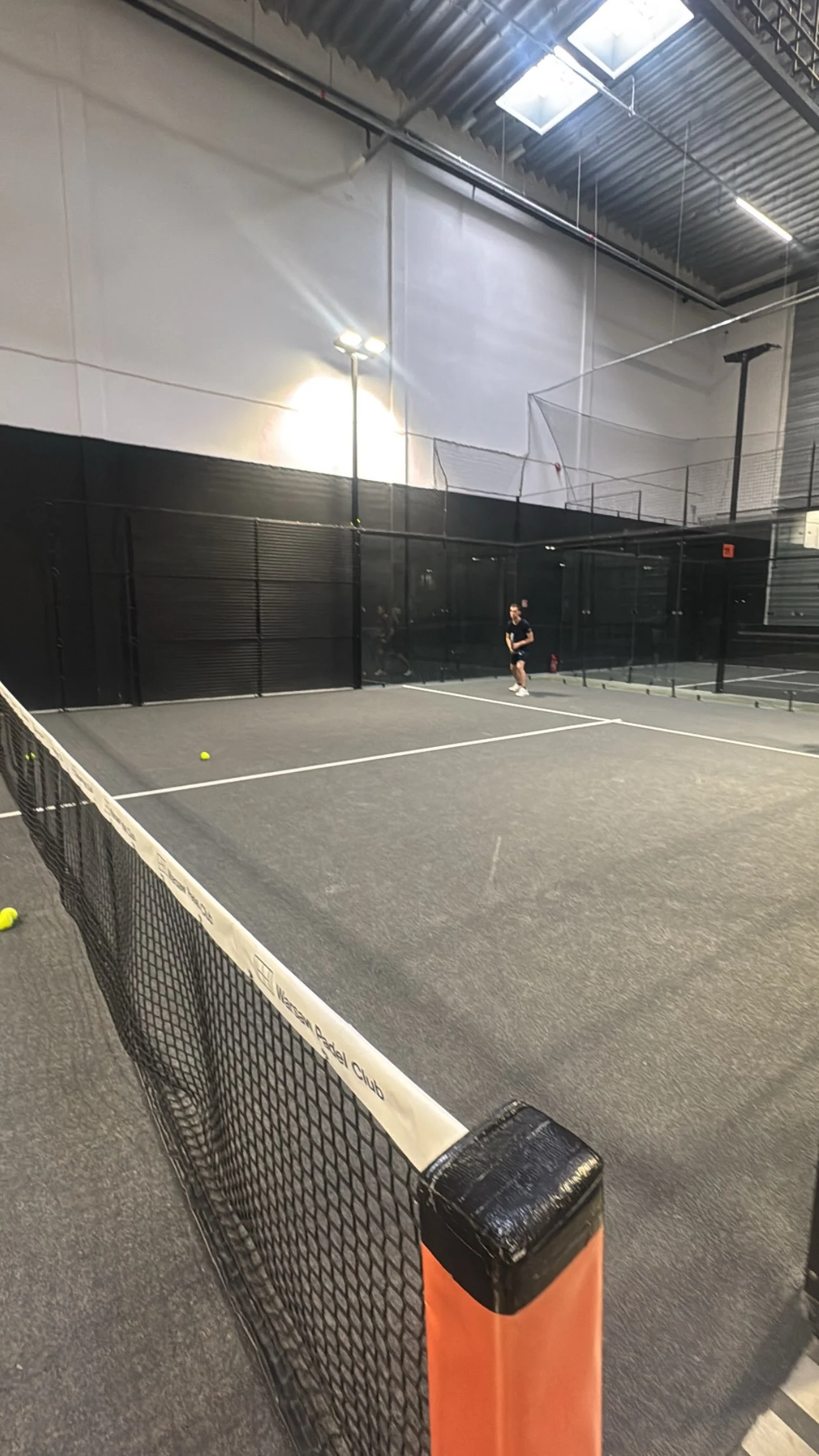 An indoor padel court seen from the net, with a player at the far end.