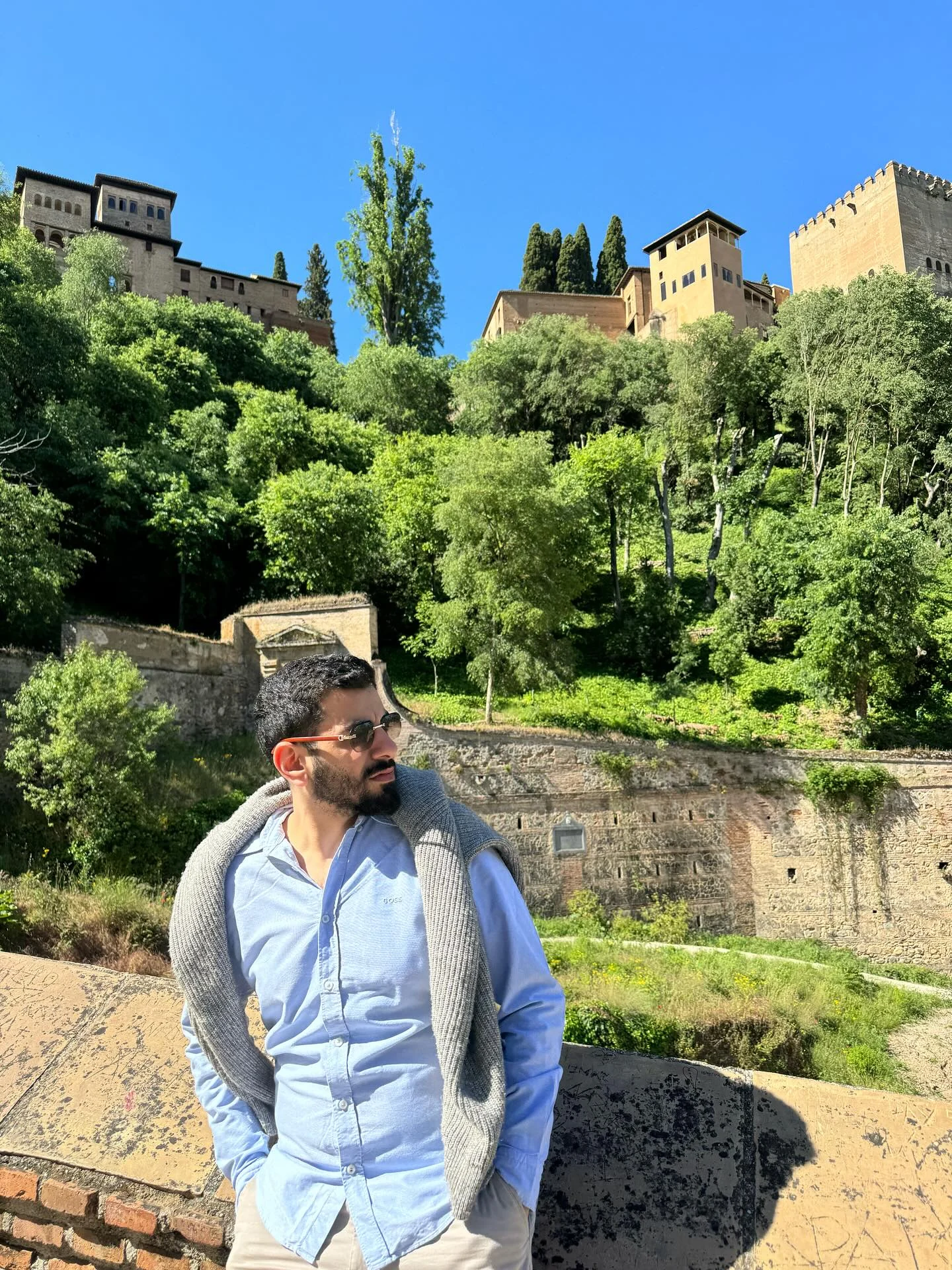 A portrait in a blue shirt below stone hilltop walls and green trees in Spain.