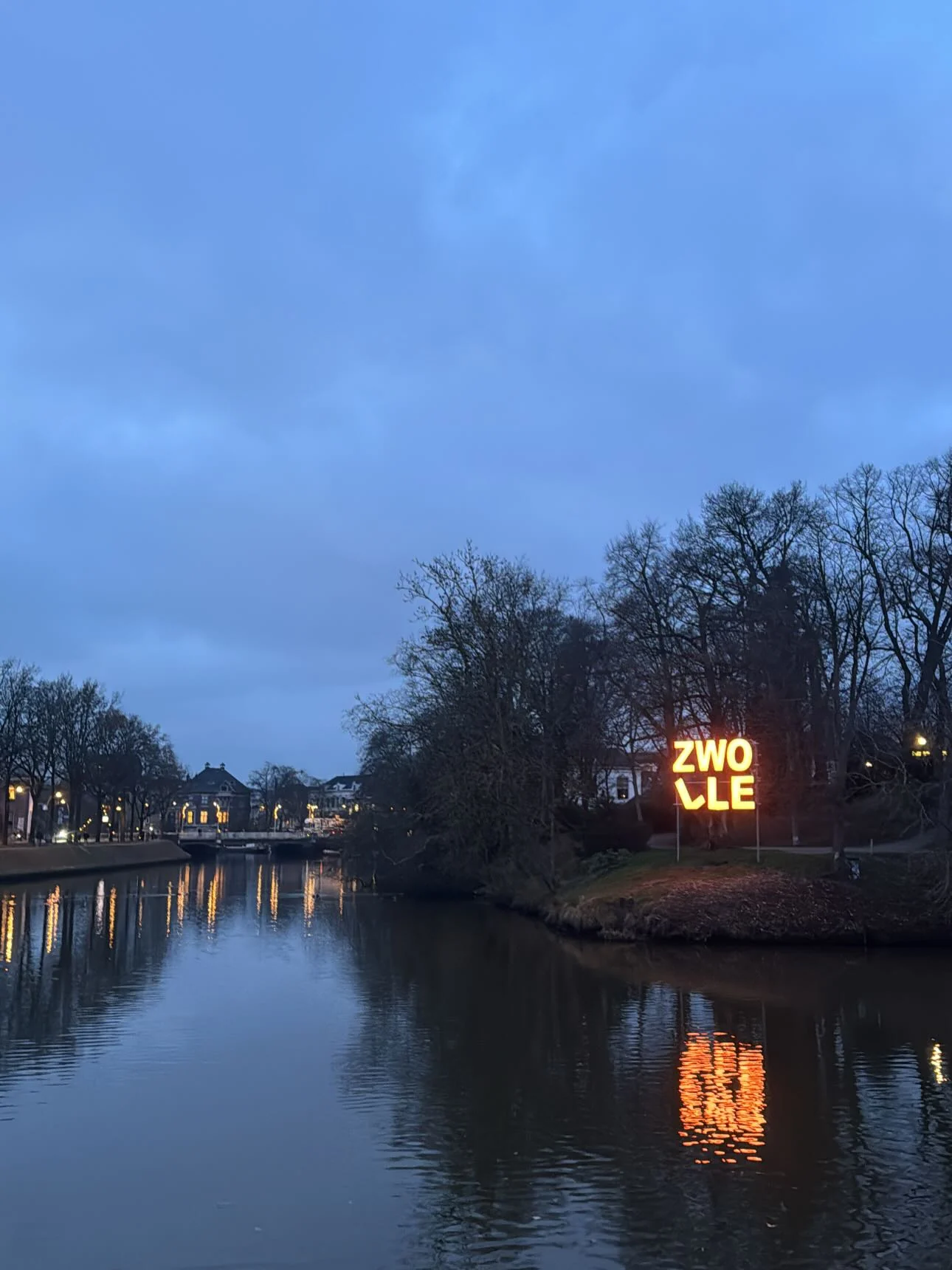 A waterfront at dusk, with bare trees and a glowing Zwolle sign reflected in the water.